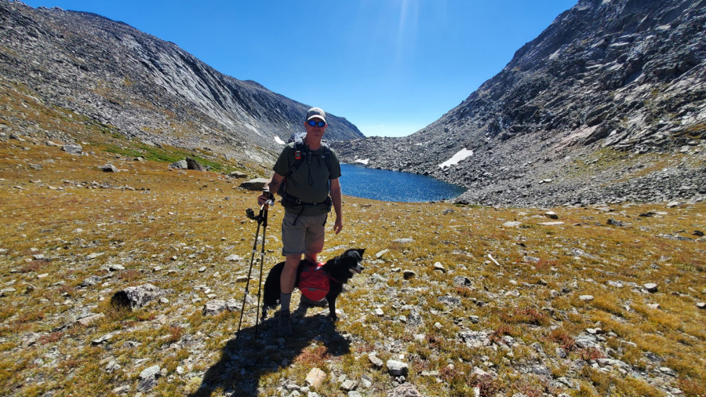 Man stands with border collie dog in an alpine setting below Hailey Pass, Wyoming, Wind River Range. He is wearing a backpack.